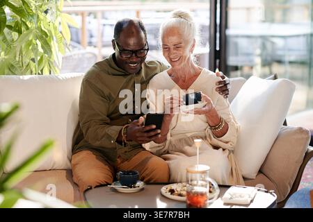 Middle-aged Black man having coffee date with senior caucasian wife sitting beside him at table in cafe Stock Photo
