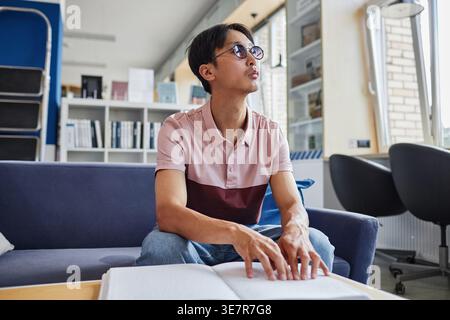 Blind Asian male student wearing eyeglasses reading book in tactile braille font sitting on couch in college library during daytime Stock Photo