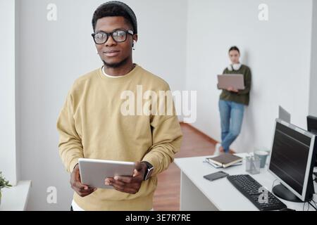 Black man wearing eyeglasses and earphone writing notes sitting at table with computer near Mixed race woman colleague leaning on wall using laptop... Stock Photo