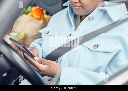 Black woman wearing seatbelt using smartphone going home after grocery shopping sitting on driver's seat in car during daytime Stock Photo