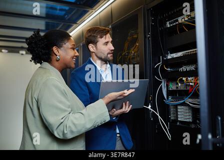 Smiling Black female IT engineer showing server cabinet to caucasian male colleague holding laptop standing in data center Stock Photo