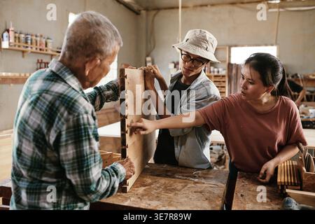 Asian male and female carpenters holding and pointing on wooden board while discussing standing at workbench in workshop Stock Photo