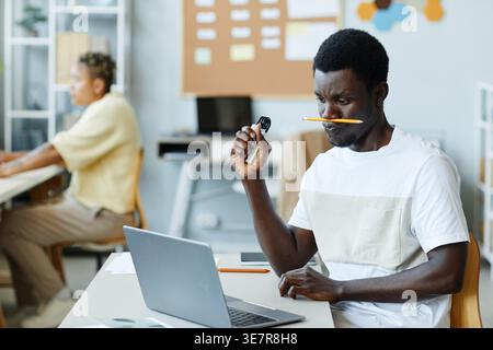 Black male IT specialist with pencil placed on nose holding wrist strengtheners while sitting at table with laptop in office Stock Photo
