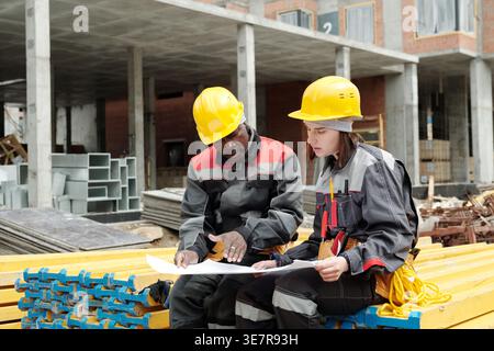Black man worker looking and discussing blueprint with caucasian female co worker while sitting on stack of  building materials in construction sit... Stock Photo