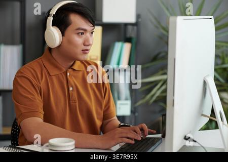 Asian male software developer wearing headphones testing new software on laptop sitting at desk in office Stock Photo