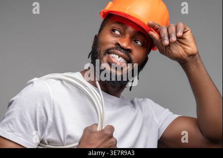 Smiling Black man wearing orange hard hat looking away with roll of wire on shoulder standing against gray background Stock Photo