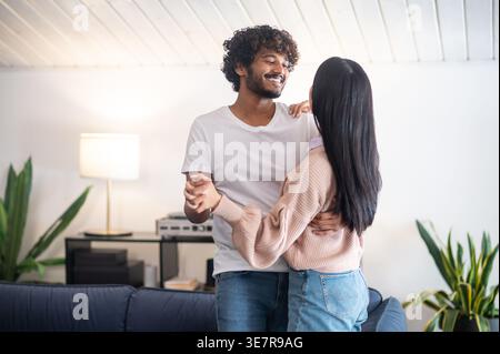 Smiling Asian couple holding hands looking at each other while standing together at home Stock Photo