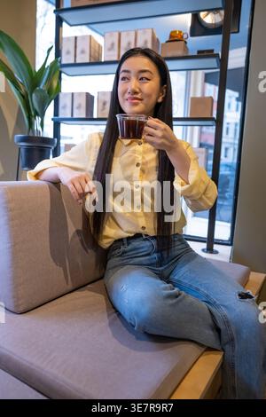 Smiling Asian woman having cup of coffee while sitting on couch in cafe Stock Photo