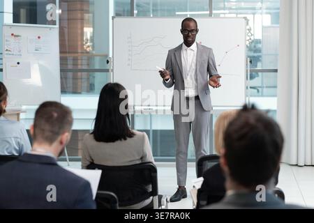 Serious Black businessman wearing eyeglasses standing and explaining graphic data on whiteboard in conference hall Stock Photo