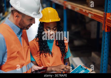 Black female worker with curly hair wearing hard hat and safety vest holding clipboard and checking inventory with caucasian male coworker while st... Stock Photo
