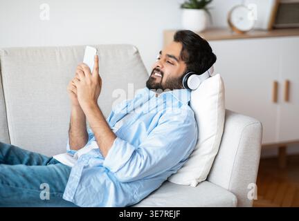 Middle Eastern man using smartphone lying on sofa in living room at home Stock Photo