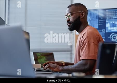 Side view portrait of Black male programmer wearing eyeglasses using computer while sitting at table with laptop in office Stock Photo