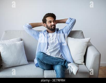 Weekend At Home. Relaxed Arab Guy Leaning Back On Couch With Hands Behind Head, Cheerful Middle Eastern Man Resting On Comfortable Sofa In Cozy Living Stock Photo