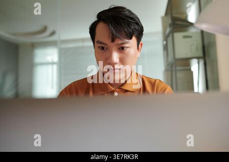 Asian male software developer working on laptop sitting at desk in office Stock Photo