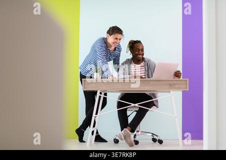 Smiling Black businessman with braids sitting at table working on laptop and having discussion with caucasian female colleague standing beside him ... Stock Photo