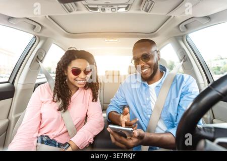 Smiling Black couple with sunglasses sharing smartphone while riding car together in city Stock Photo