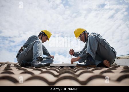 Asian male builders in hardhats in uniform installing solar panels on roof of client's house  during the daytime Stock Photo