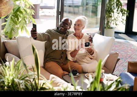 Middle-aged Black man taking selfie with senior caucasian wife sitting beside him at table during coffee date in cafe Stock Photo