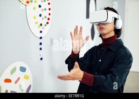 Asian man wearing VR headset experiencing virtual reality while standing in art gallery Stock Photo