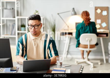 Serious Asian male programmer wearing eyeglasses using laptop sitting at table with computer near Black female colleague in office Stock Photo