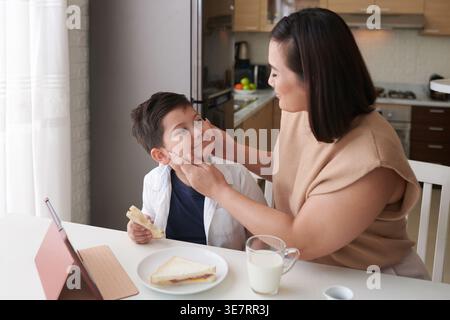Smiling Asian mother squishing cheeks of Mixed race son while eating sandwiches in kitchen at home during daytime Stock Photo