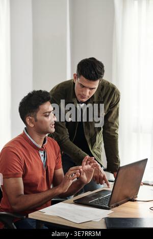 Indian male programmer standing and pointing on table discussing computer program with male colleague in office Stock Photo