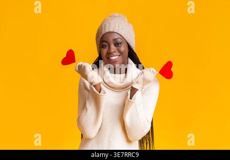 Black woman wearing knitted hat and gloves holding red hearts on sticks standing against yellow background in studio Stock Photo
