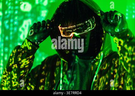 Serious Black man wearing led augmented reality glasses adjusting hoodie while standing in dark room with green neon lights Stock Photo