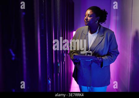 Serious Black female IT engineer inspecting server network holding tablet standing in dark room with purple neon lights in data center Stock Photo