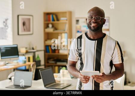 Black man wearing eyeglasses holding tablet standing near desk with laptops in office Stock Photo