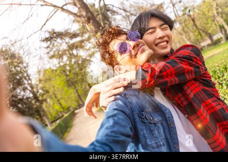 Asian and caucasian gay couple taking selfie while embracing each other standing in park during daytime Stock Photo
