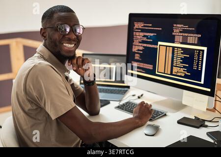 Black male programmer with hand on chin wearing eyeglasses sitting at table with laptop and computer in office Stock Photo