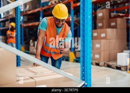 Black male worker wearing hard hat and safety vest scanning boxes using barcode scanner during work in warehouse Stock Photo
