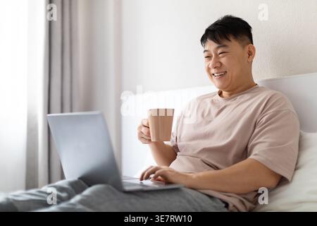 Relaxed happy handsome middle aged korean man wearing pajamas drinking aromatic fresh morning coffee and using modern pc laptop in bed, surfing on Int Stock Photo