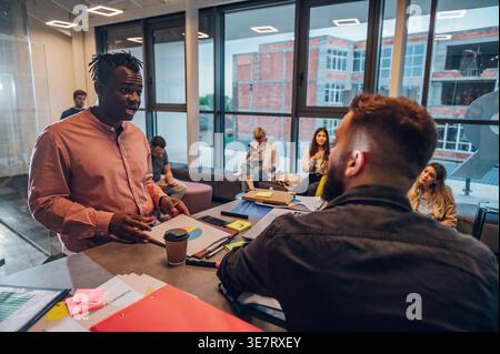 Black man standing while discussing plans with caucasian male coworker sitting across table with documents in office Stock Photo
