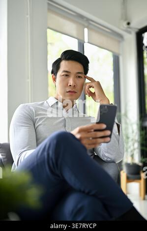 Serious Asian man touching head using smartphone sitting with crossed legs on chair in office during daytime Stock Photo