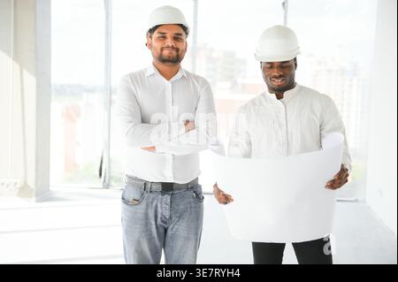 Black male engineer holding blueprint standing with Indian male colleague crossing arms during work on construction site Stock Photo