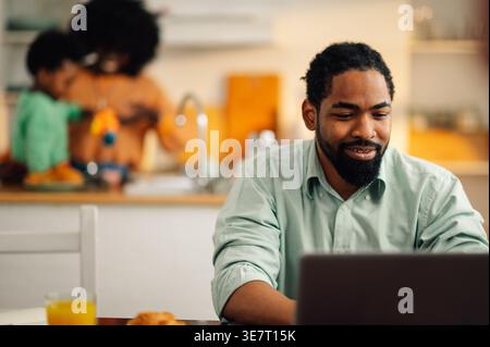Smiling Black father sitting at table working on laptop near wife and son playing at kitchen counter at home Stock Photo