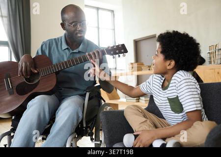 Smiling Disabled Black father in glasses sitting in wheelchair playing guitar together with son at home Stock Photo