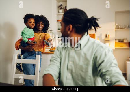 Cheerful Black mother with afro holding her son standing in kitchen near husband sitting at table and looking at them at home Stock Photo