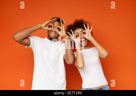 Cheerful Black couple showing okay symbol around eyes while standing against orange background in studio Stock Photo