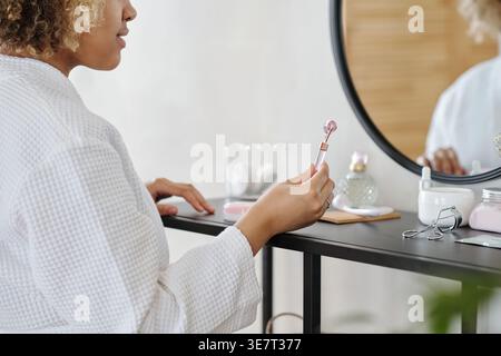 Black woman in bathrobe holding facial roller standing in front of mirror in bathroom Stock Photo