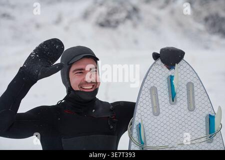 Smiling Mixed race man in surfing suit holding surf board and waving his hand while standing on snowy mountain during winter Stock Photo
