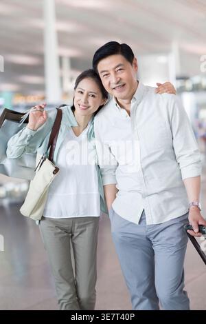 Smiling middle-aged Chinese couple standing close with their luggage in the airport during flight Stock Photo