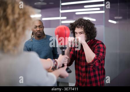 Focused mixed-race man doing boxing with white female coworker holding punching ball standing near Black male colleague in office Stock Photo