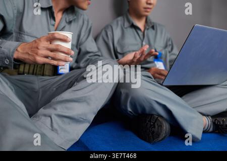 Asian male builders in uniform holding cup of coffee sharing laptop discussing construction plan sitting with crossed legs against gray wall on con... Stock Photo