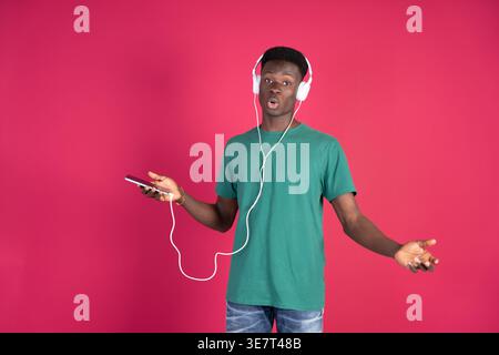 Black man in green shirt wearing headphones connected to smartphone singing while standing against pink background in studio Stock Photo