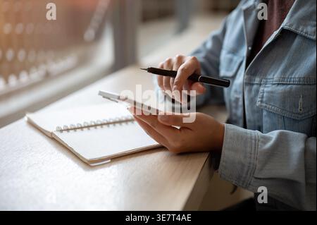 Cropped image of Thai woman in denim jacket browsing on smartphone and holding stylus pen while sitting at table with notebook indoors Stock Photo