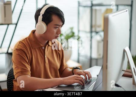 Asian male software developer wearing headphones testing new software on laptop sitting at desk in office Stock Photo