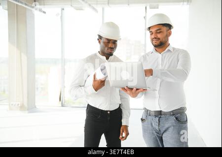 Diverse group of male engineers wearing hard hats holding blueprint and laptop discussing project during work on construction site Stock Photo
